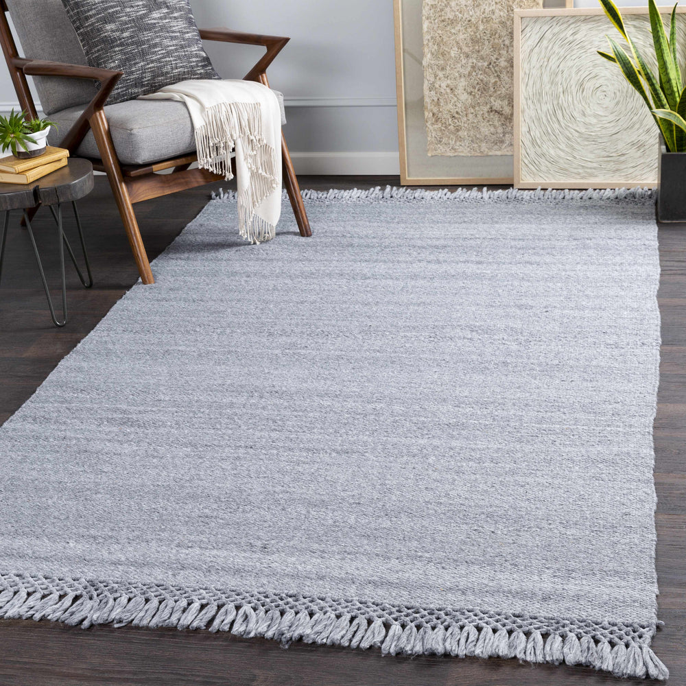 Gray hand-woven area rug with fringed edges, shown on a floor with furniture